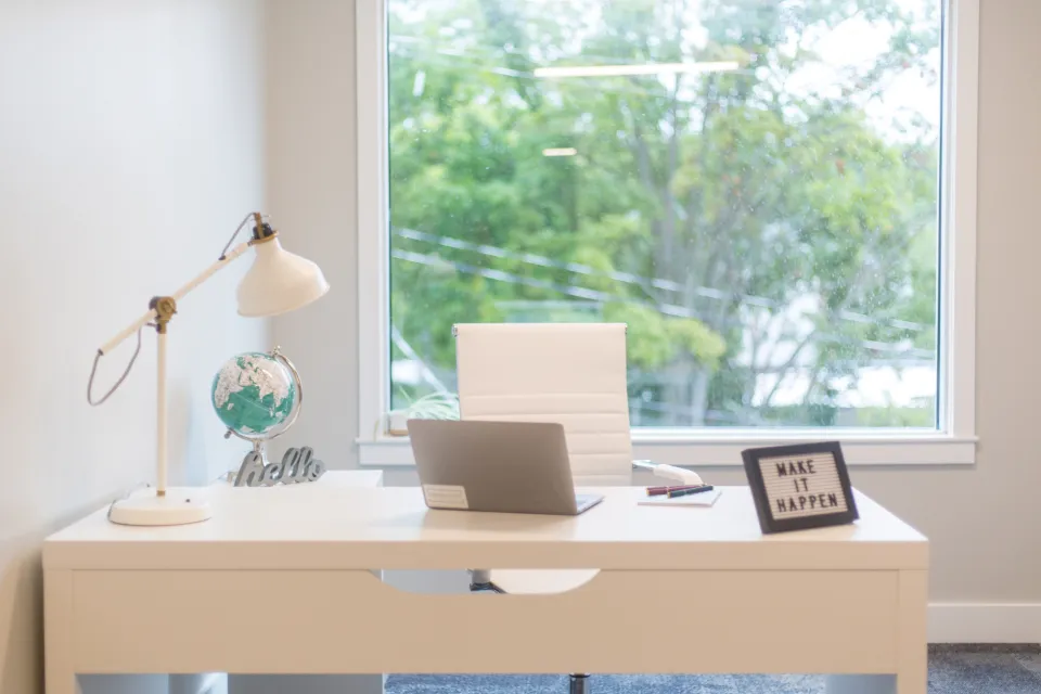 Bright home office with a white desk, laptop, desk lamp and an empty chair by a window