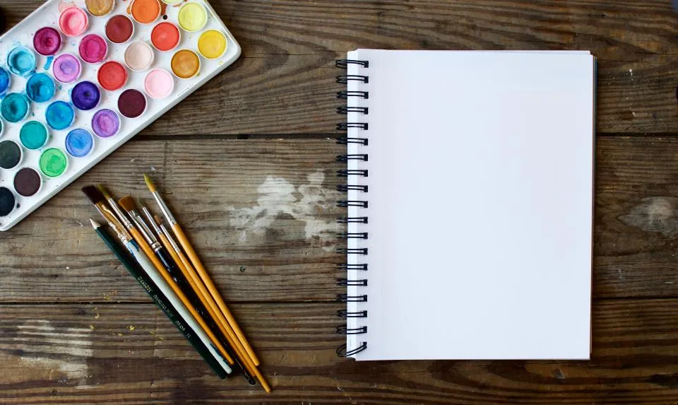 Watercolour paint set, brushes and a blank sketchbook on a wooden table