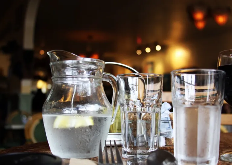 Glass pitcher of water with lemon slices next to glasses of water on a table