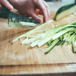 Hands slicing courgette into strips on a wooden chopping board