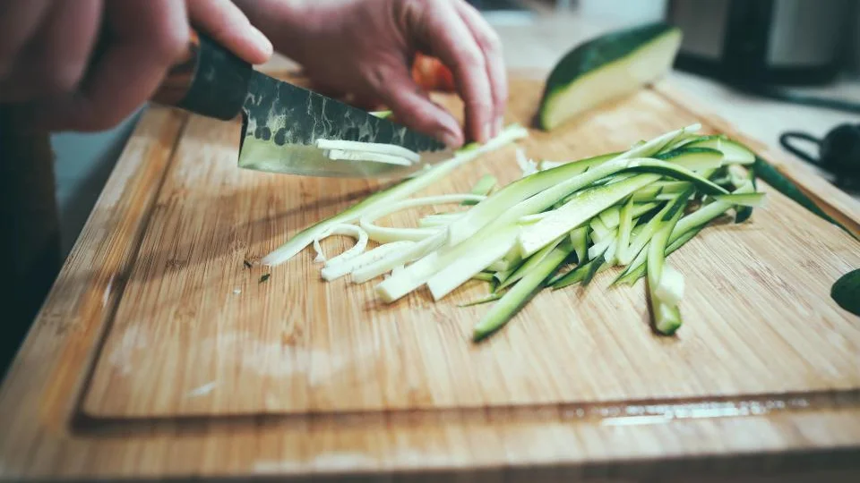 Hands slicing courgette into strips on a wooden chopping board